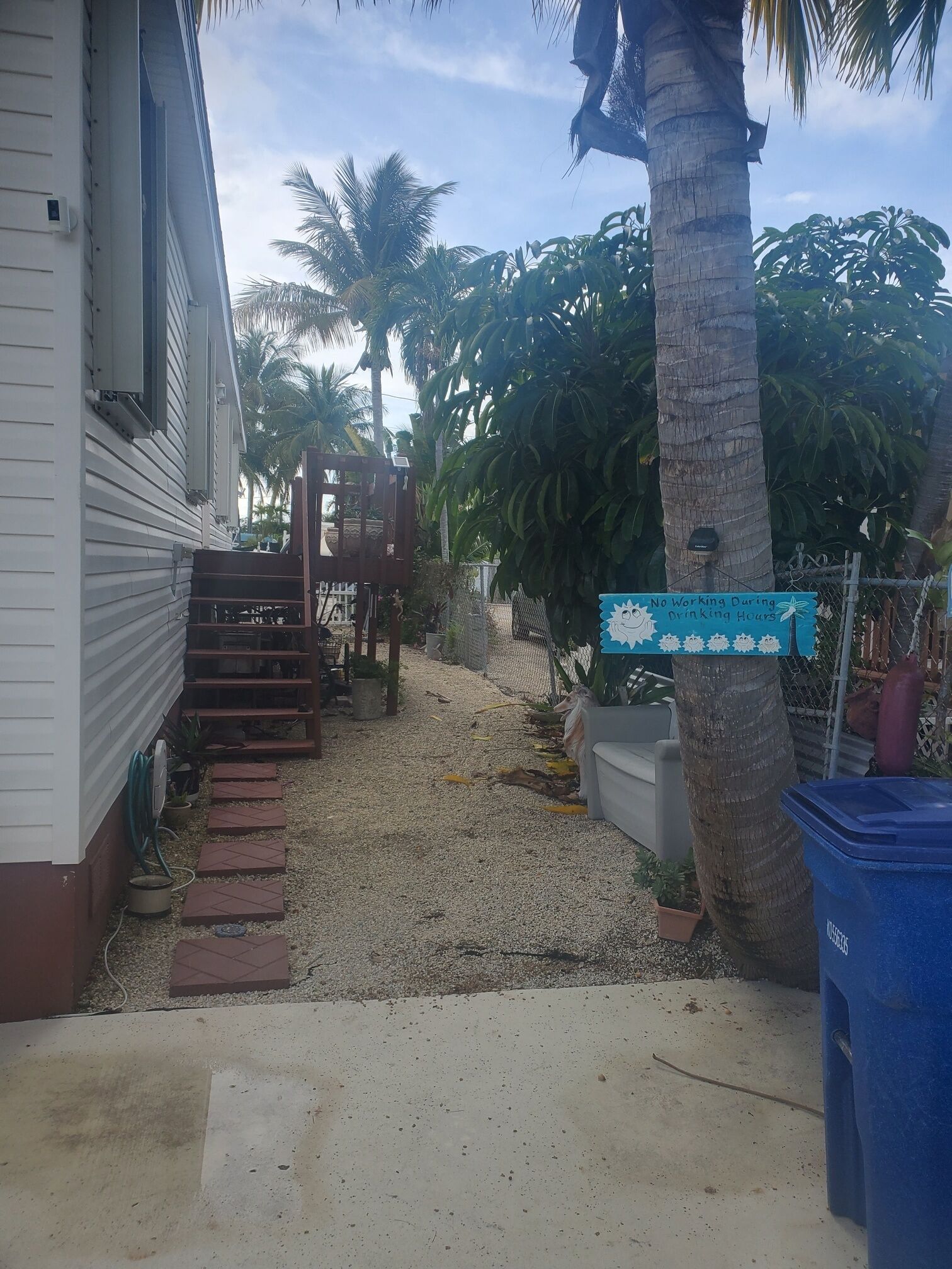 A residential area with a house, a gravel pathway, palm trees, and a sign indicating no working during drinking hours.