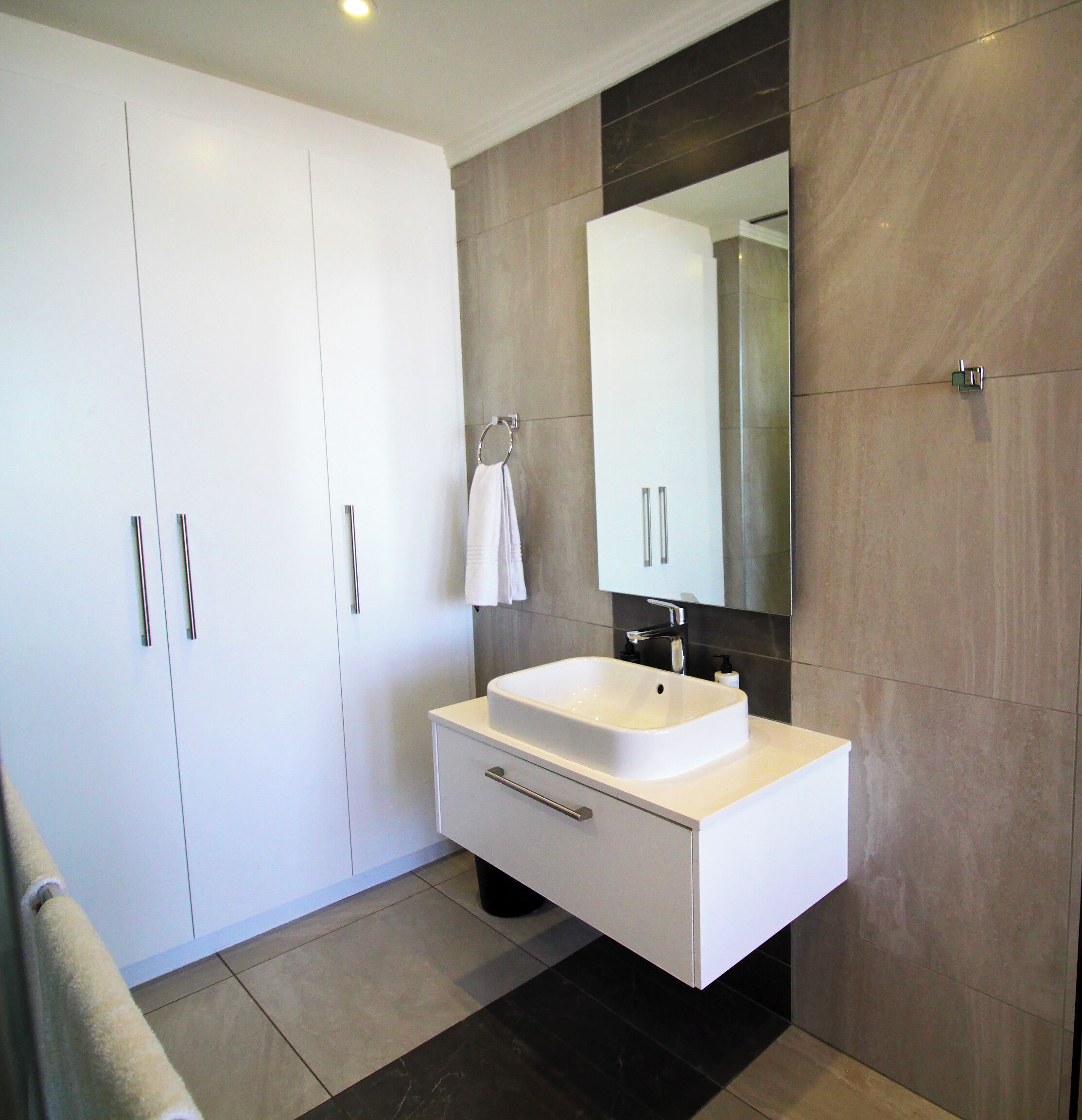 Une salle de bain moderne avec un meuble-lavabo blanc, un grand miroir et un mur recouvert de carreaux foncés.