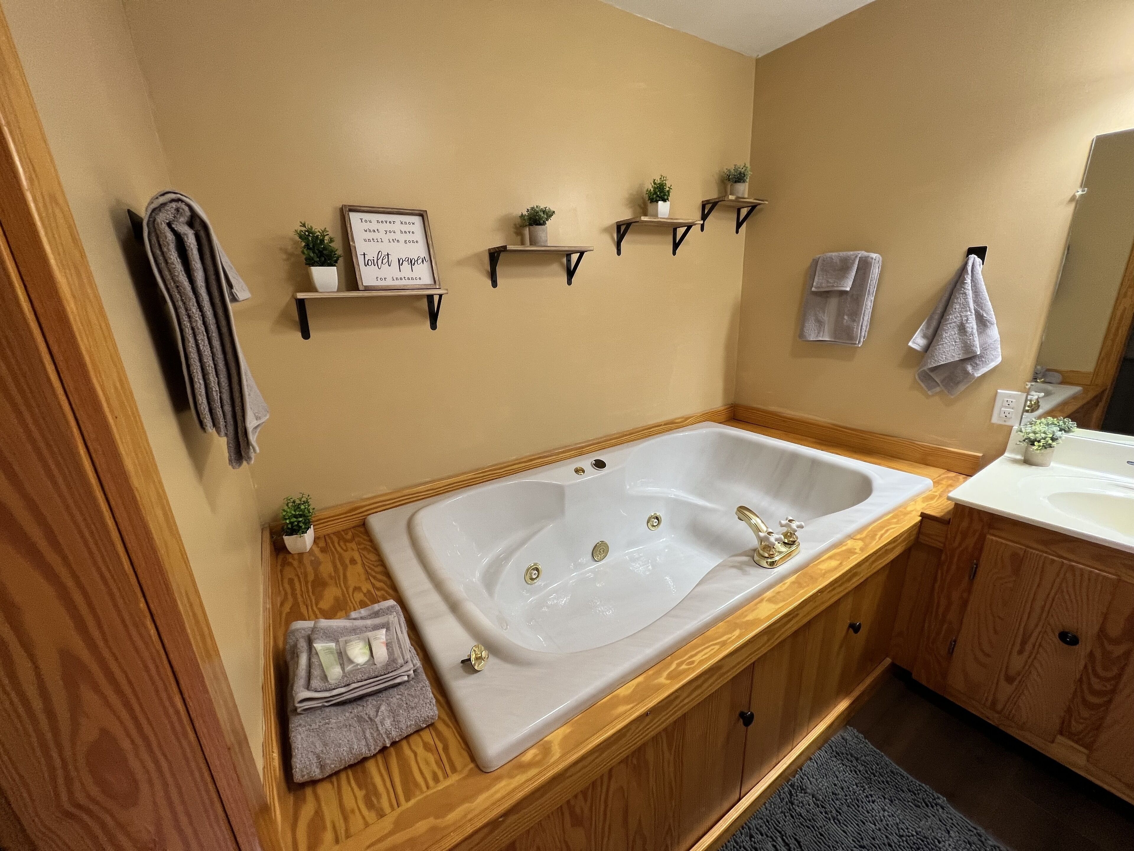 Une salle de bain avec une grande baignoire, des meubles en bois, un meuble-lavabo avec un évier et un miroir. On y trouve des serviettes et une citation encadrée accrochée au mur.