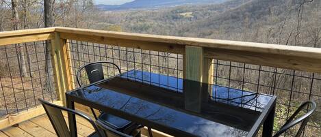 A wooden deck with a glass table and metal chairs overlooking a mountainous landscape.