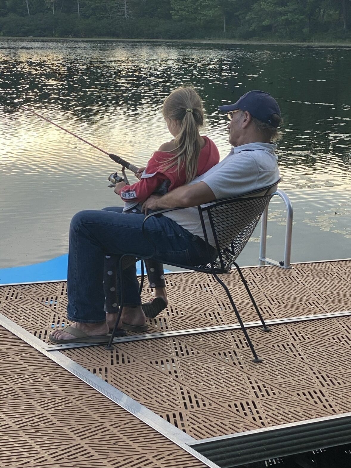 Two people fishing on a dock with a calm lake in the background.