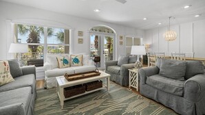 A spacious living room with a white ceiling fan, grey sofas, a coffee table with a woven tray, and large windows with blinds.