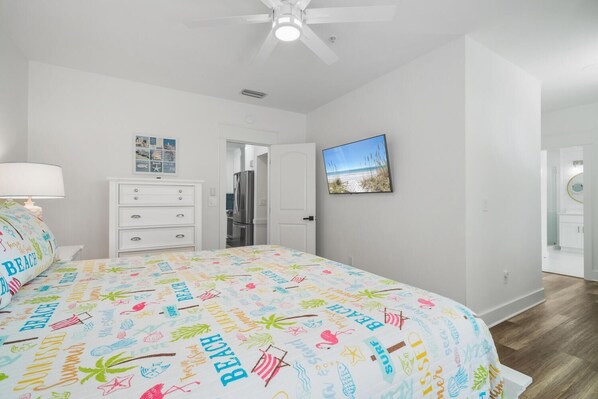 A bedroom with a bed, a dresser, a ceiling fan, a refrigerator, a door leading to another room, and a framed picture on the wall.