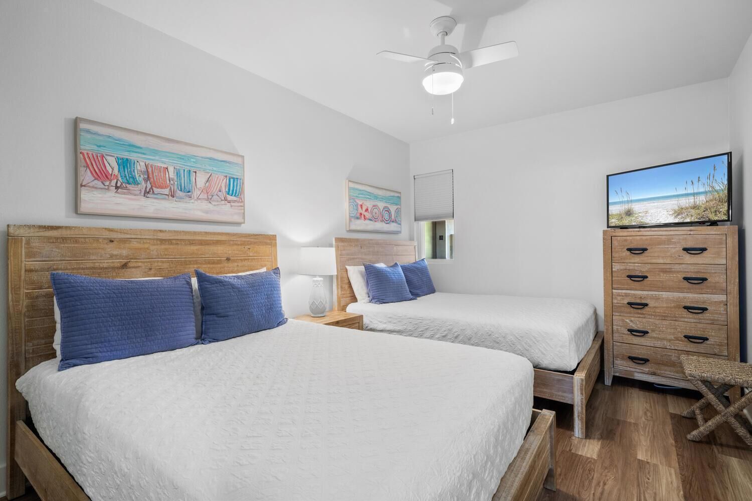 A hotel room with two beds, wooden headboards, blue pillows, a wooden dresser, a television, and artwork on the walls.