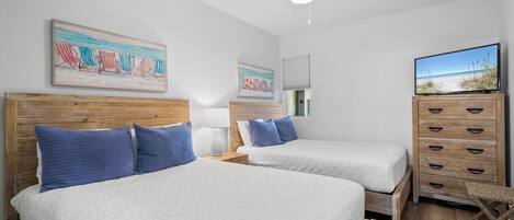 A hotel room with two beds, wooden headboards, blue pillows, a wooden dresser, a television, and artwork on the walls.