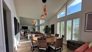 Een ruime eetruimte met een grote houten tafel en rieten stoelen, grote ramen met zonwering en een moderne kroonluchter.