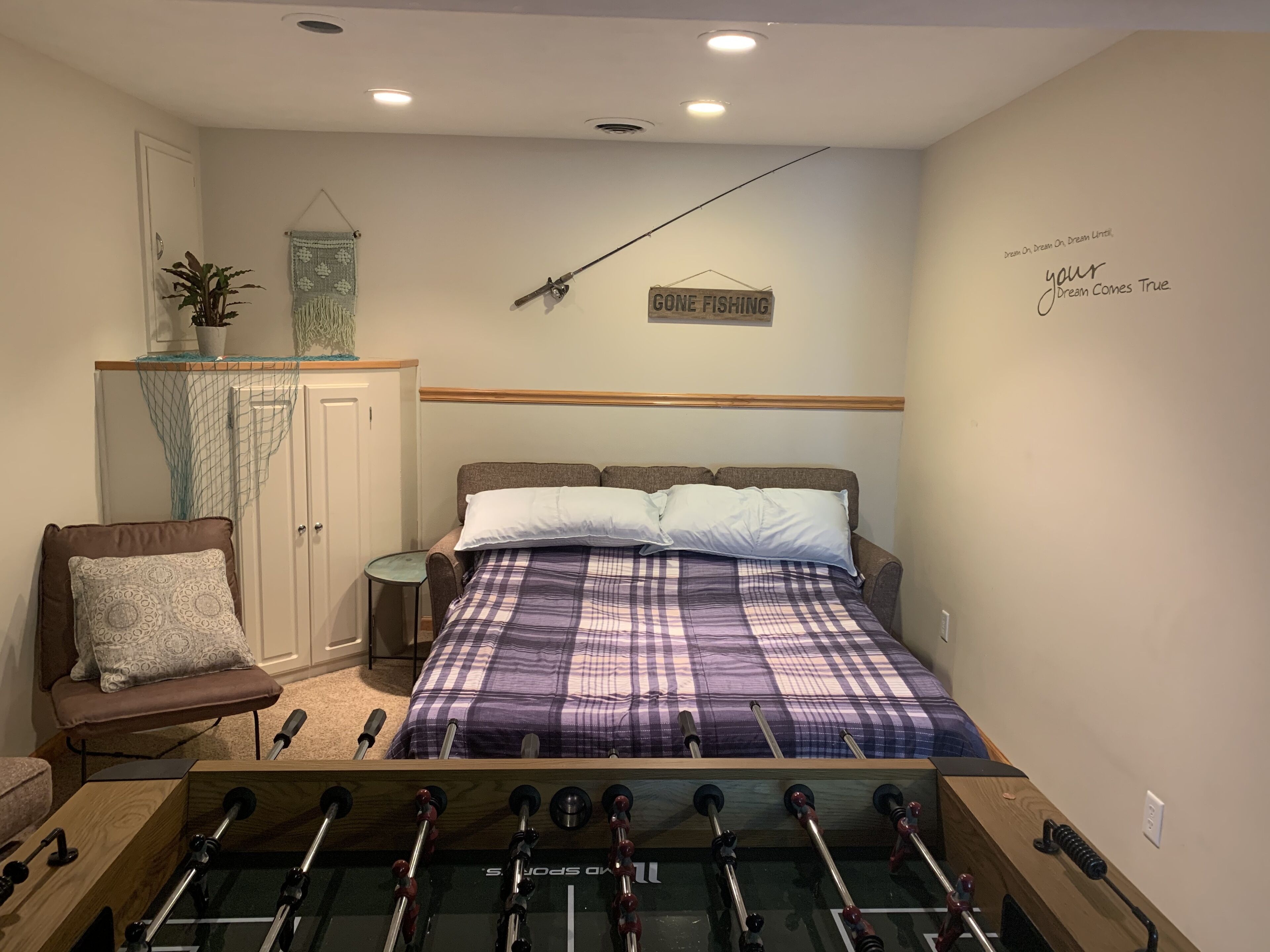 Une chambre avec un lit, une chaise, un baby-foot, une petite table et un panneau décoratif au mur.