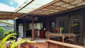 A covered outdoor area with a wooden deck, a dining table with chairs, and a kitchen area visible through an open door. - Bungalow Temanea Pirae Tahiti (Papeete)