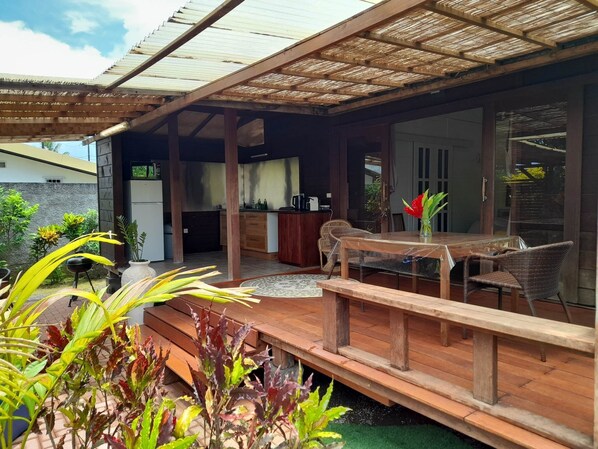 A covered outdoor area with a wooden deck, a dining table with chairs, and a kitchen area visible through an open door.