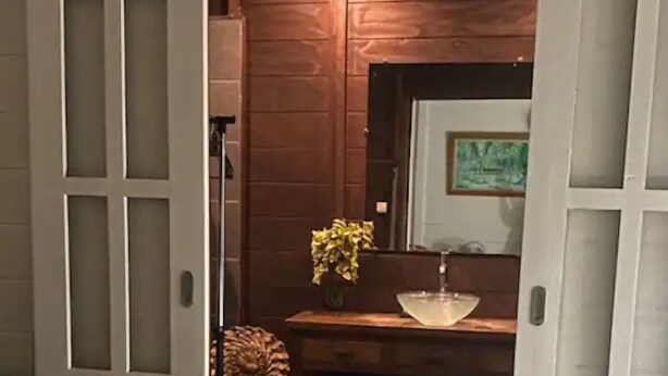 A bedroom with a wooden floor, a bed with a white cover, a wooden dresser, a mirror, and a small plant on a shelf. - Bungalow Temanea Pirae Tahiti (Papeete)