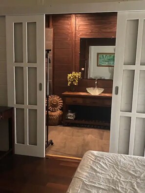 A bedroom with a wooden floor, a bed with a white cover, a wooden dresser, a mirror, and a small plant on a shelf.