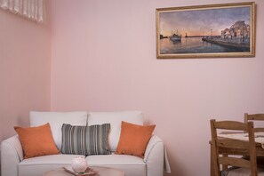 A living room with a white sofa, two orange cushions, a striped pillow, and a small table with a decorative item. A framed painting of a waterfront scene hangs on the wall.