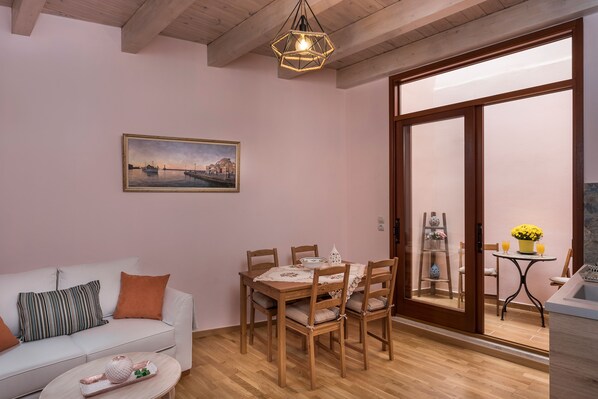A cozy living room with a white sofa, a wooden dining table with chairs, a painting on the wall, and a sliding glass door leading to a balcony with a bathtub and a small table with flowers.