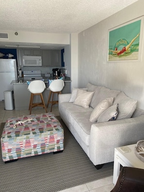 A modern living room with a grey sofa, a patterned ottoman, and a small table with a decorative item. A kitchen with white cabinets and a microwave is visible in the background. - Beachfront piece of heaven! (Daytona Beach Shores)