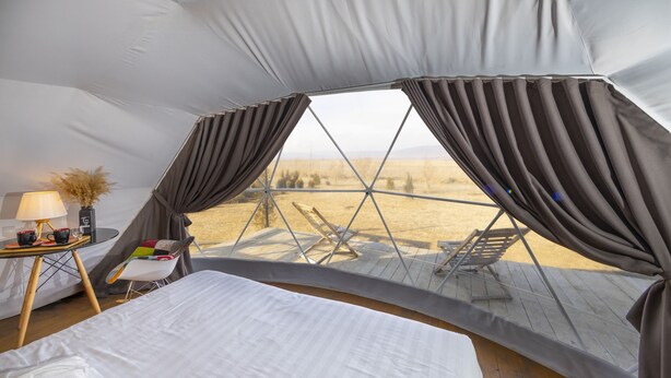 A tent with a bed, a table with a lamp, and a view of the outdoors.