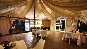 A tented dining area with tables and chairs, wooden flooring, and string lights.