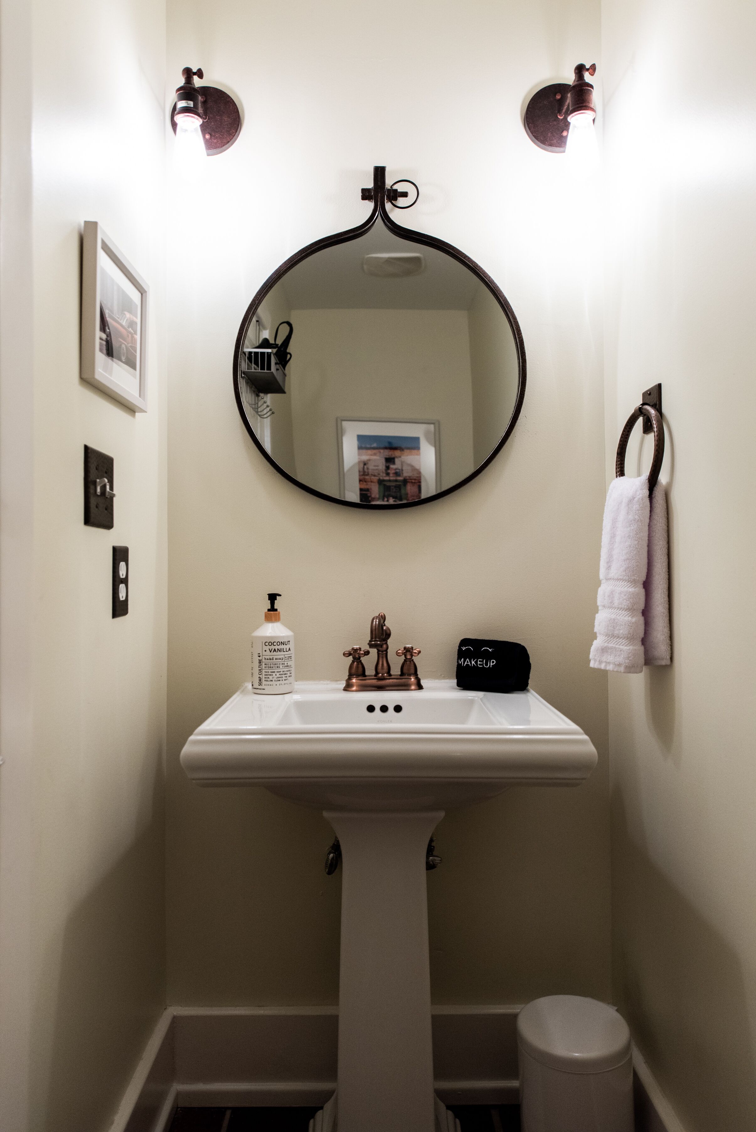Une salle de bain avec un lavabo sur colonne, un miroir rond, un porte-serviettes et une petite étagère sur laquelle se trouvent un flacon et une trousse de maquillage.