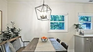 A dining area with a wooden table, chairs, a hanging light fixture, and a window with a view of trees.
