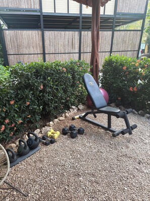 An outdoor workout area with a bench, kettlebells, and dumbbells on a gravel surface, surrounded by greenery and a wooden fence.