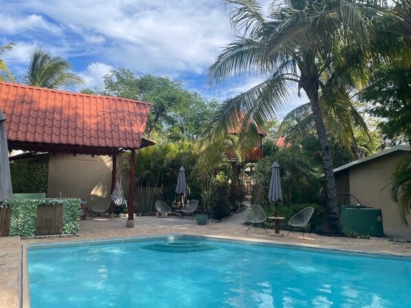 A swimming pool with lounge chairs and umbrellas, surrounded by palm trees and a building with a red roof.