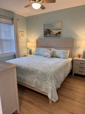 A bedroom with a bed, a ceiling fan, a nightstand, a lamp, a picture on the wall, and a window with blinds.