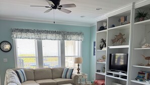 A living room with a sectional sofa, a coffee table, a TV on a stand, a ceiling fan, and a bookshelf. - Oceanfront - The Ocean Club - Sand, Sun & Family Fun - Indian Beach/Emerald Isle (Indian Beach)
