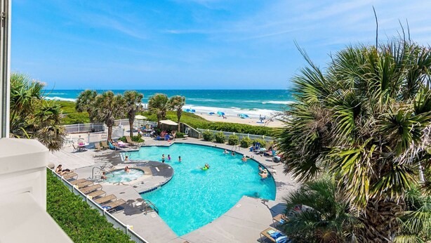 A pool area with a view of the beach, surrounded by palm trees and lounge chairs. - Oceanfront - The Ocean Club - Sand, Sun & Family Fun - Indian Beach/Emerald Isle (Indian Beach)