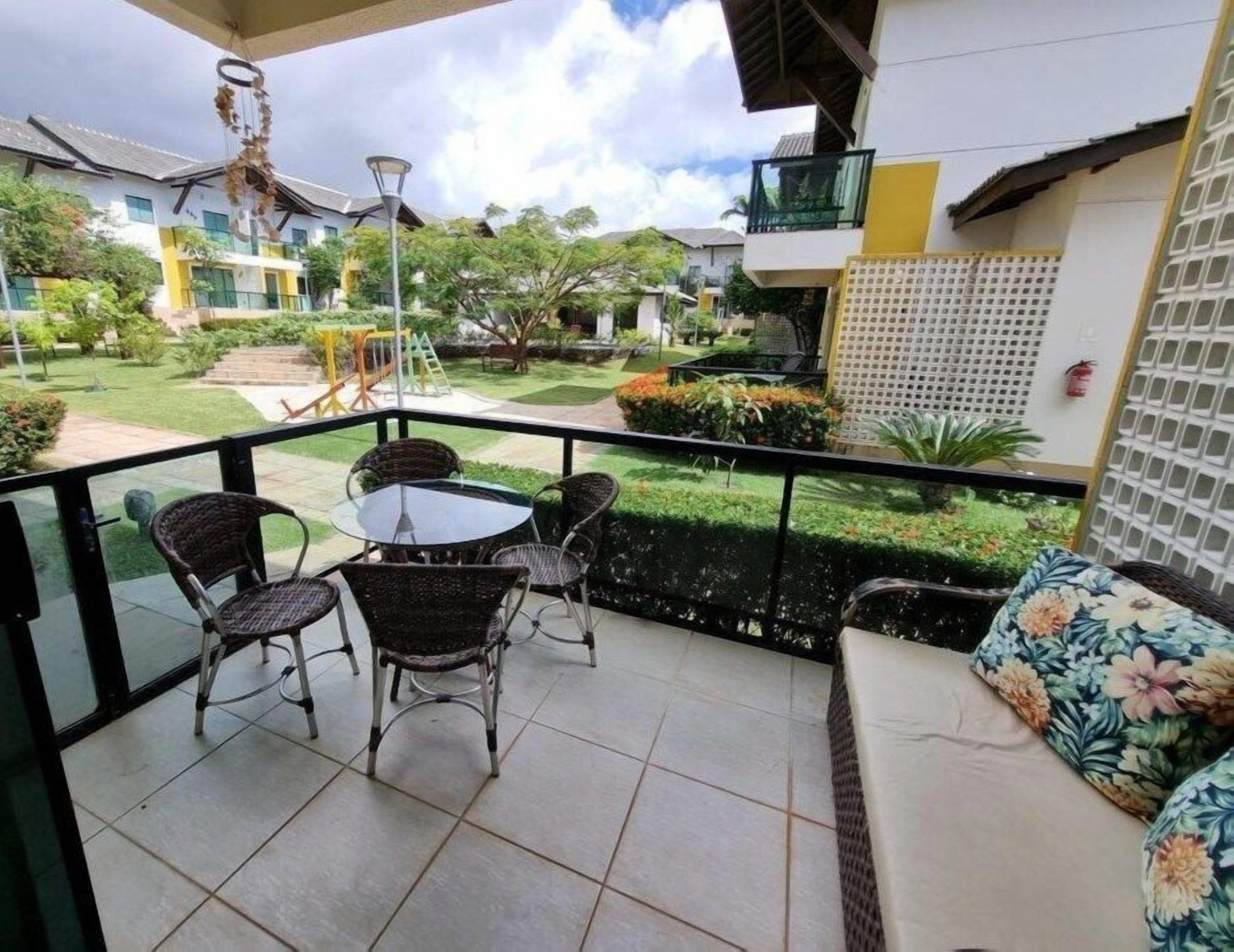 A balcony with wicker chairs and a table, overlooking a well-maintained garden and neighboring buildings.