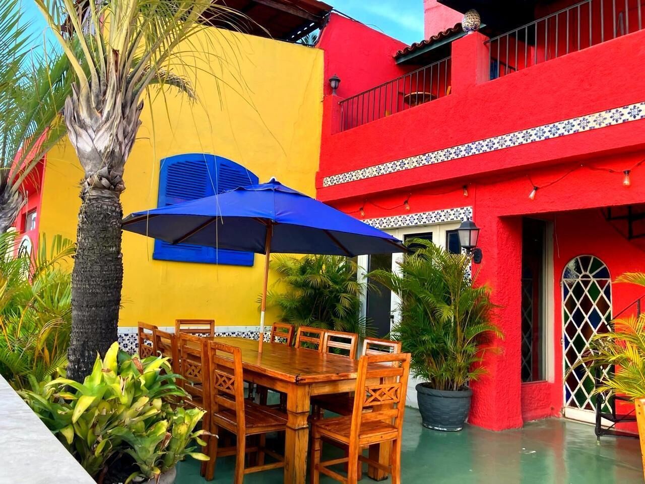 A wooden dining table with chairs, a blue umbrella, and potted plants in an outdoor setting.