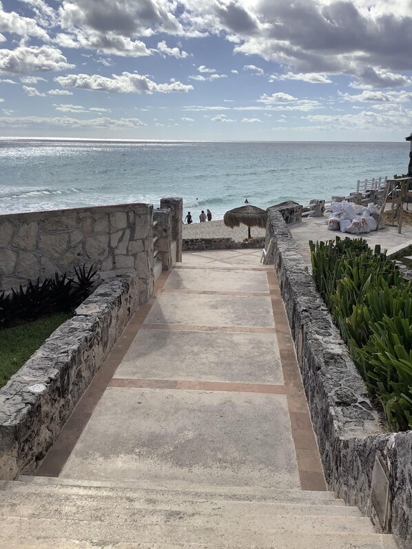 A stone pathway leading to a beach with a view of the ocean and a thatched umbrella.