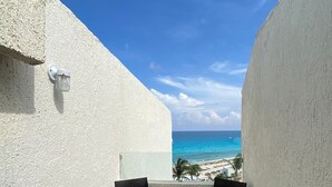 Un balcone con due sedie nere e un tavolino, con vista su una spiaggia e acqua cristallina, con alcune palme.