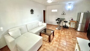 Ein Wohnzimmer mit einem weißen Sofa, einem Couchtisch, einem Deckenventilator, einer Küchenzeile mit Kühlschrank und einem kleinen Barhocker.