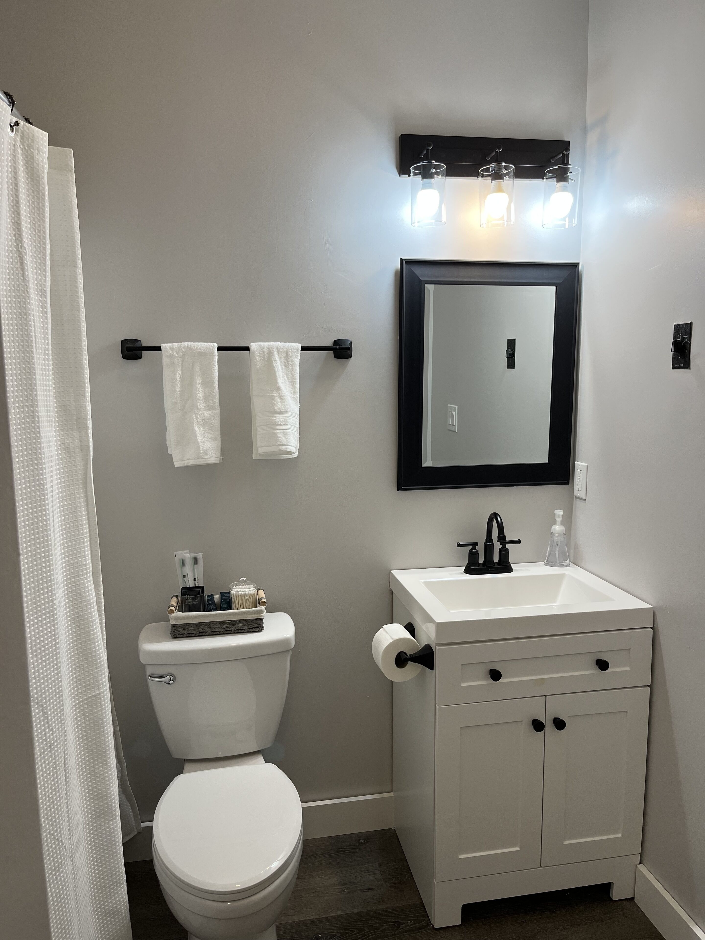 Un baño con inodoro, lavamanos y espejo. Hay toallas colgadas en un perchero y una cortina de ducha.