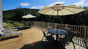 Eine Holzterrasse mit zwei runden Tischen, jeweils mit Sonnenschirm, umgeben von Stühlen.