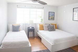 Un dormitorio con dos camas, una ventana con persianas, un ventilador de techo y una pequeña mesa con lámpara.