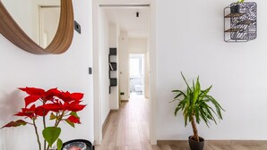 A hallway with a wooden floor, a potted plant, a mirror, and a wall-mounted light fixture.