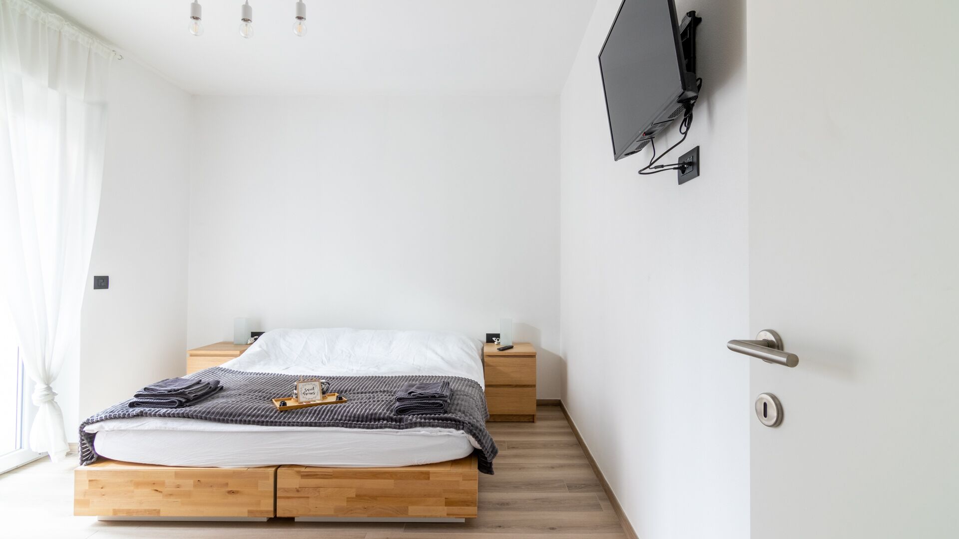Une chambre à coucher au style minimaliste, avec un lit en bois, des tables de chevet, un téléviseur fixé au mur et une fenêtre avec des rideaux.