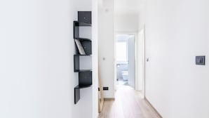 A hallway with a black wall-mounted shelf unit, a wooden broom leaning against the wall, and a door leading to a bathroom.