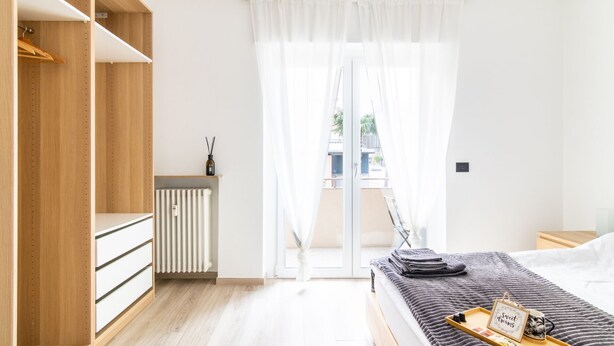 A modern bedroom with a wooden wardrobe, a radiator, a bed with a tray and a picture frame, and a view through a glass door.