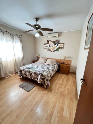 A bedroom with a bed, a ceiling fan, a nightstand, a wall-mounted air conditioner, and a painting.