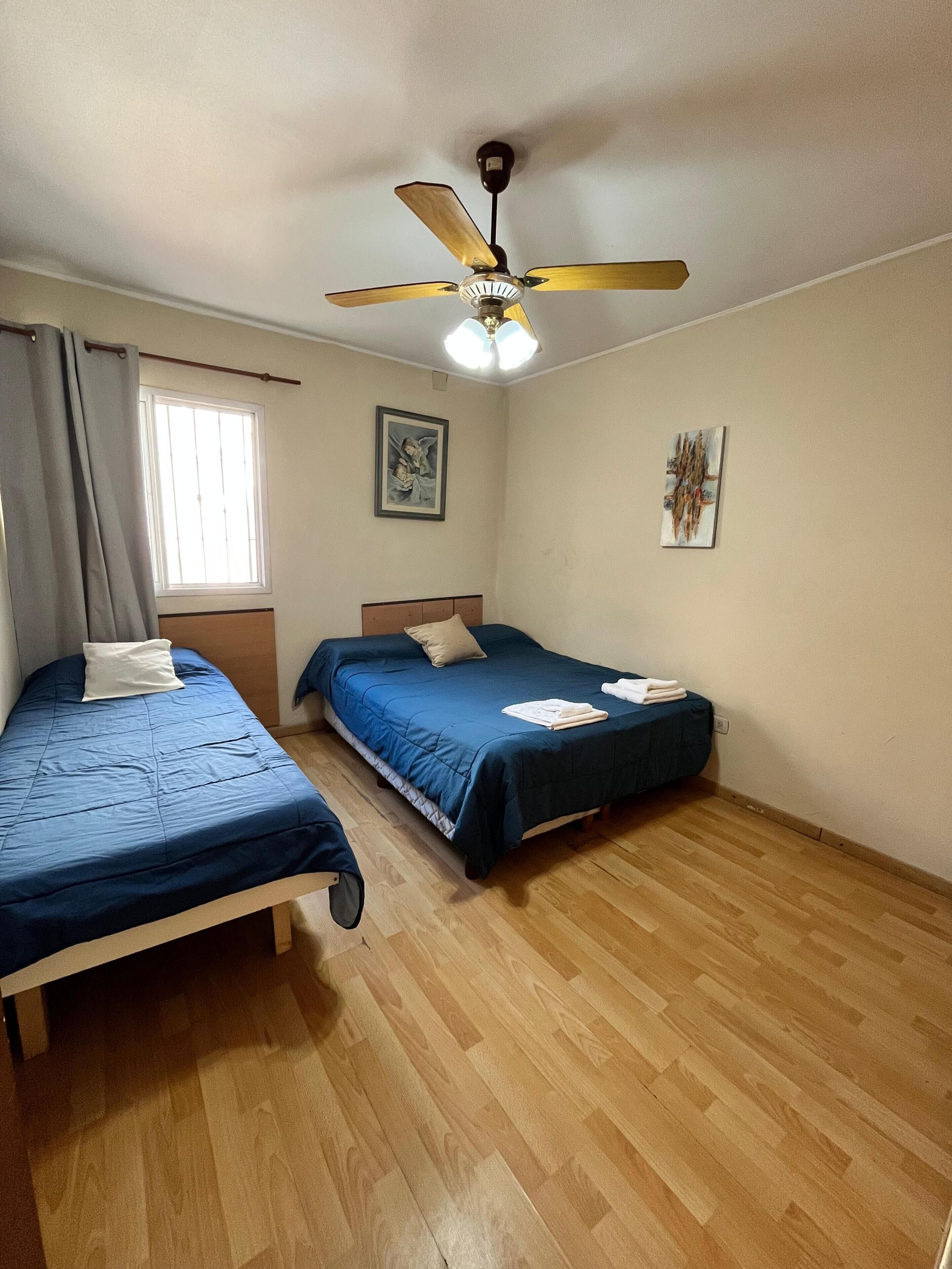 A room with two single beds, a ceiling fan, a window with curtains, and a wooden floor.