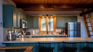 A kitchen with teal cabinets, a stainless steel refrigerator, a microwave, and a large window. There are three black bar stools and a wooden bench. - Modern Mountain Chalet Near Storyland & Summer Fun (Intervale)