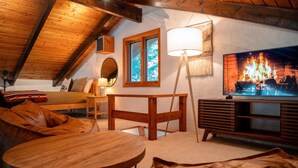A cozy loft bedroom with a wooden ceiling, a TV displaying a fireplace, a wooden table, and a comfortable bed with pillows. - Modern Mountain Chalet Near Storyland & Summer Fun (Intervale)