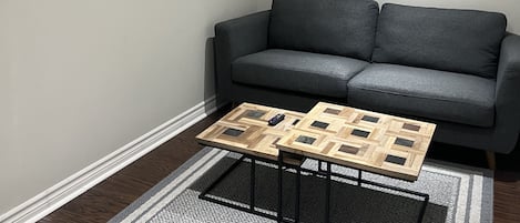 Una sala de estar con un sofá gris, una mesa de centro de madera, un tapete estampado, un reloj de pared y un cartel enmarcado.
