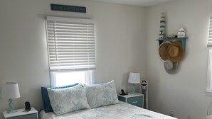 A bedroom with a bed, two nightstands, a window with blinds, a wall-mounted hat rack, and a sign above the window.