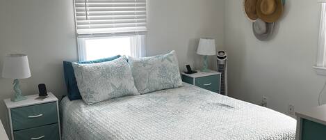 A bedroom with a bed, two nightstands, a window with blinds, a wall-mounted hat rack, and a sign above the window.