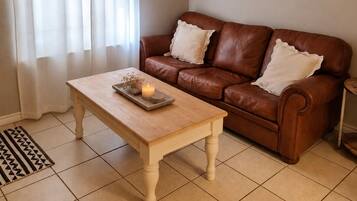 A living room with a brown leather sofa, a wooden coffee table with decorative items, a ceiling fan, and a window with sheer curtains.