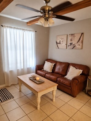 A living room with a brown leather sofa, a wooden coffee table with decorative items, a ceiling fan, and a window with sheer curtains.