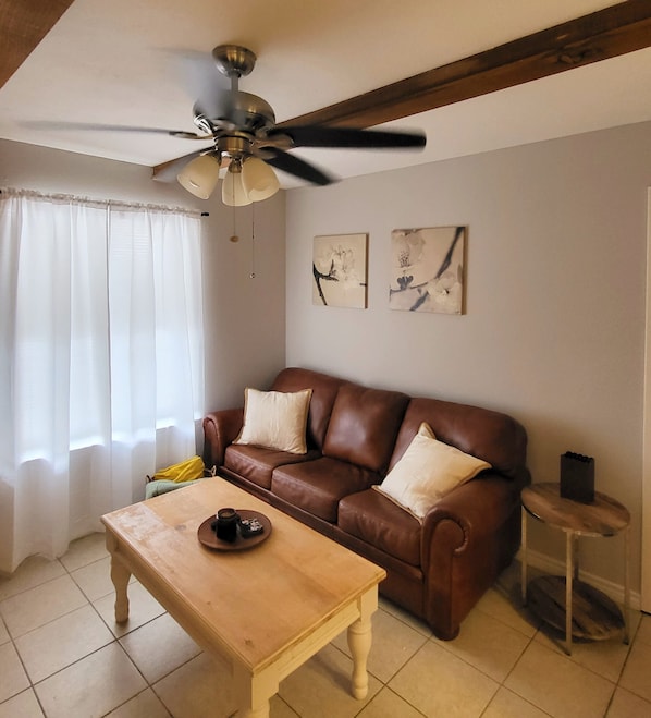 Una sala de estar con un sofá de cuero marrón, una mesa de centro de madera, un ventilador de techo y dos cuadros colgados en la pared.