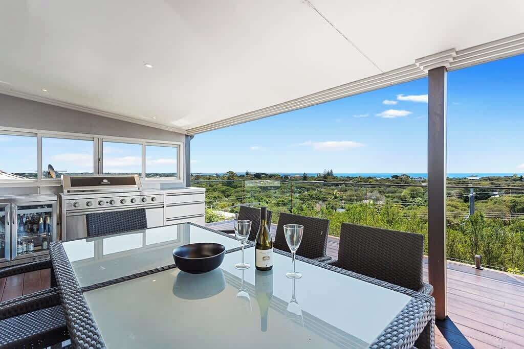 Een eethoek buiten met een glazen tafel, rieten stoelen en een barbecue.